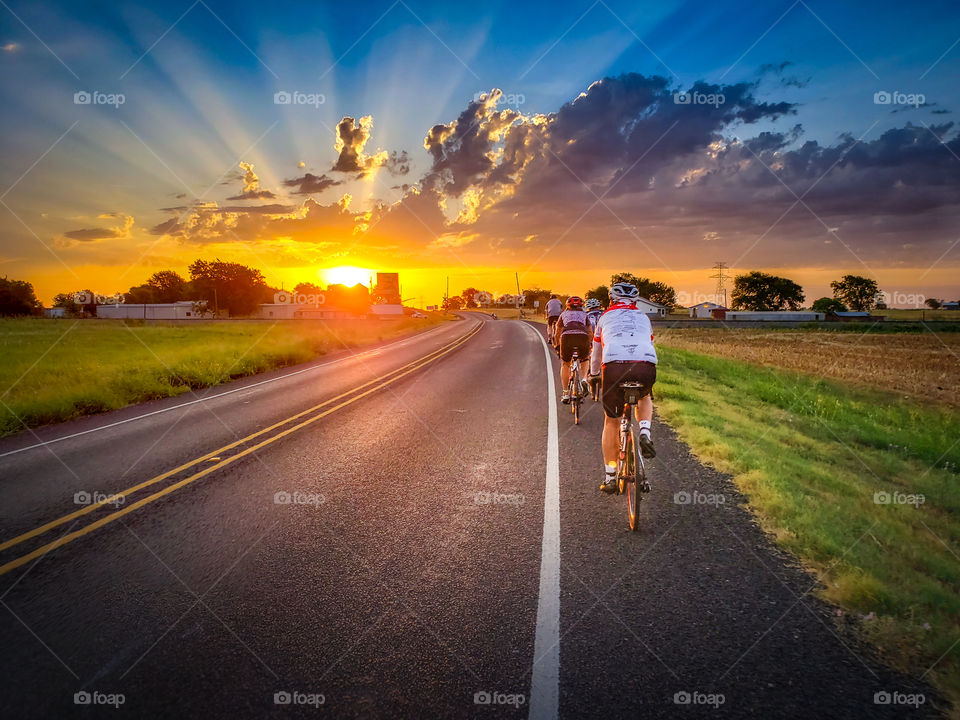 A long line of cyclists riding along a rural highway, into a beautiful warm sunrise.