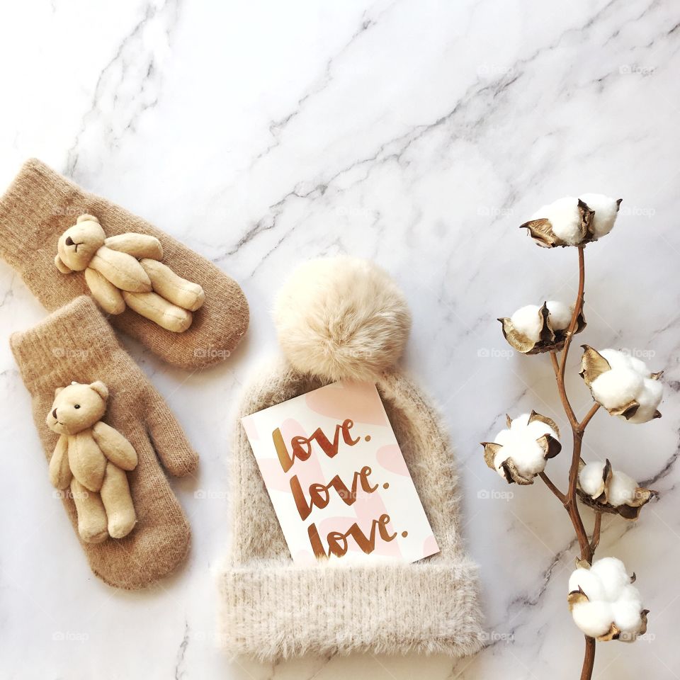 Flat lay items. Winter hat, card with inscription love, cotton flower, mittens lying on marble background 