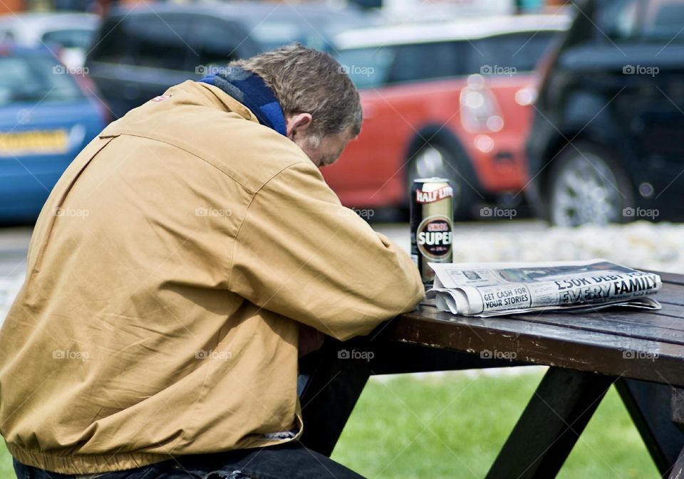 A man sits with his head down on a bench drink cheap lager with a newspaper which has depressing headlines 