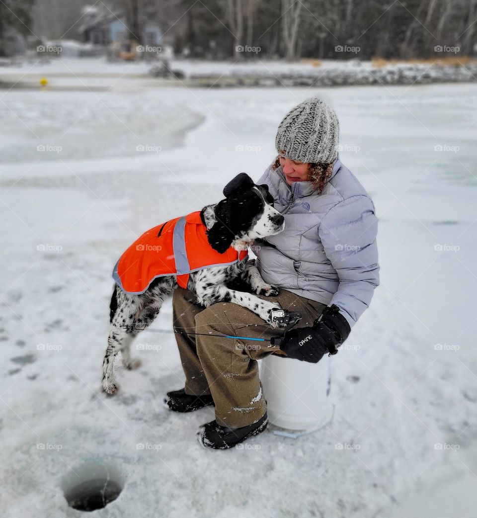 Ice fishing, woman sitting on a bucket fishing with her dog on her lap.