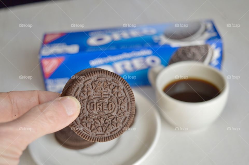 A persons fingers holding an Oreo cookie and a cup of coffee in the background