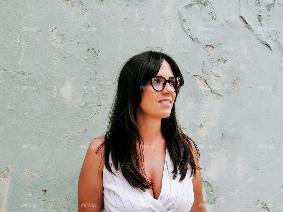 Portrait of beautiful young woman wearing sundress and eyeglasses, standing in front of weathered wall