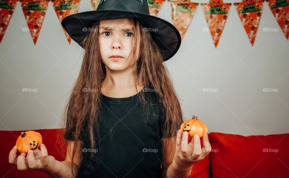 Portrait of a beautiful little caucasian girl in a witch costume and hat with a sad emotion on her face holds two small decorative pumpkins on the background of a white wall with holiday flags and an orange sofa, vtd side close-up. Concept of sad kid