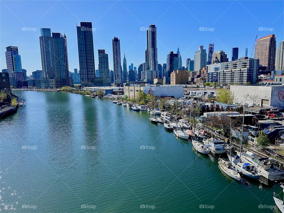 This is “Newtown Creek” with its many boats seen from the “Pulaski Bridge” that connects LIC, Queens to “Greenpoint”, Bklyn. Across the water in the distance we see “Manhattan” including the “Empire State Building”. 2024. Hypnotic Productions
