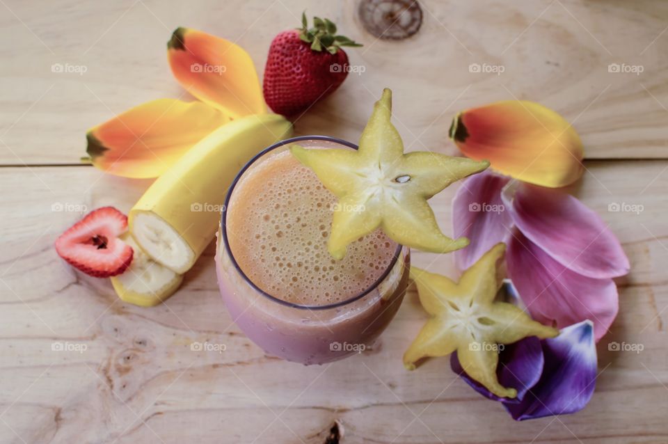 Beautiful smoothie background high angle view of smoothie with thick bubbles and fresh tropical fruit strawberry, star fruit, banana on wood table with colorful flower petals 