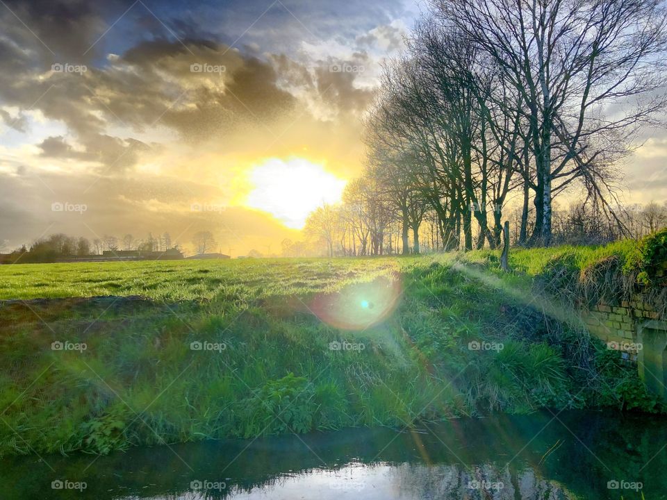 Golden sunset in a dark grey Cloudy sky over a grassland landscape with a small river running through it lined with bare trees