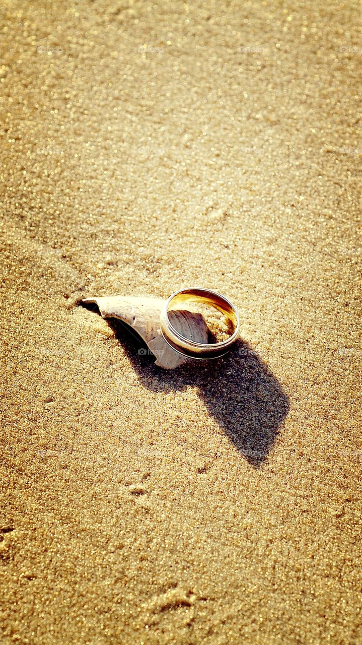 A wedding band sits atop a seashell in the sand.