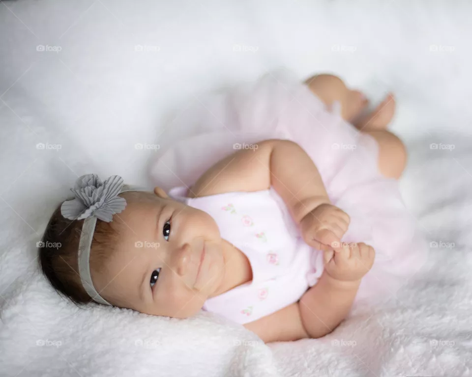 Soft photo of a baby lying down on a white fluffy blanket, wearing a pink dress with a tutu and gray bow in her hair. A gentle smile completes the sweet feeling of love she has.