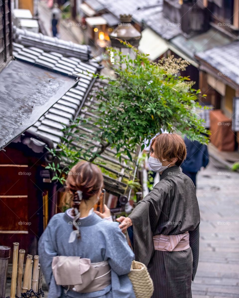 A young man holds his hand out in a gesture to invite his girlfriend. Posing for picture as the girl snaps a photo of him on her smartphone. Both wearing yukata, a summer style kimono. Traditional Japanese clothes. Ninenzaka and Sannenzaka district.