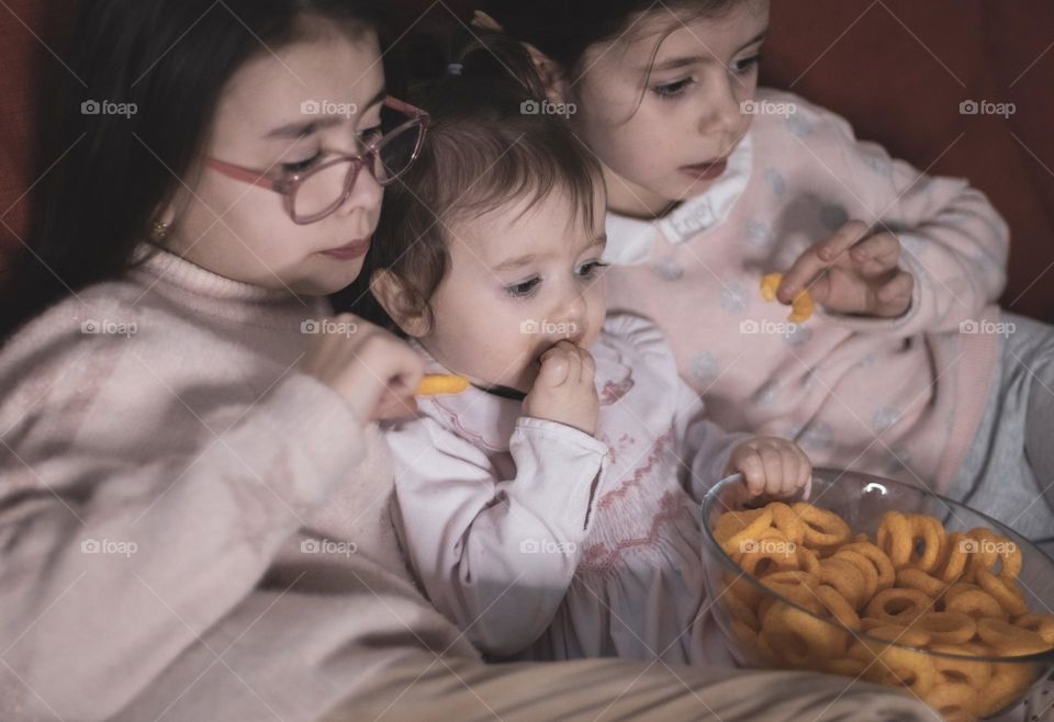 Portrait Three beautiful caucasian girls sisters eat corn rings with a transparent glass dish and enthusiastically with real emotions watch cartoon on a tablet while sitting together on a sofa, close-up side view. Family watching movie concept.