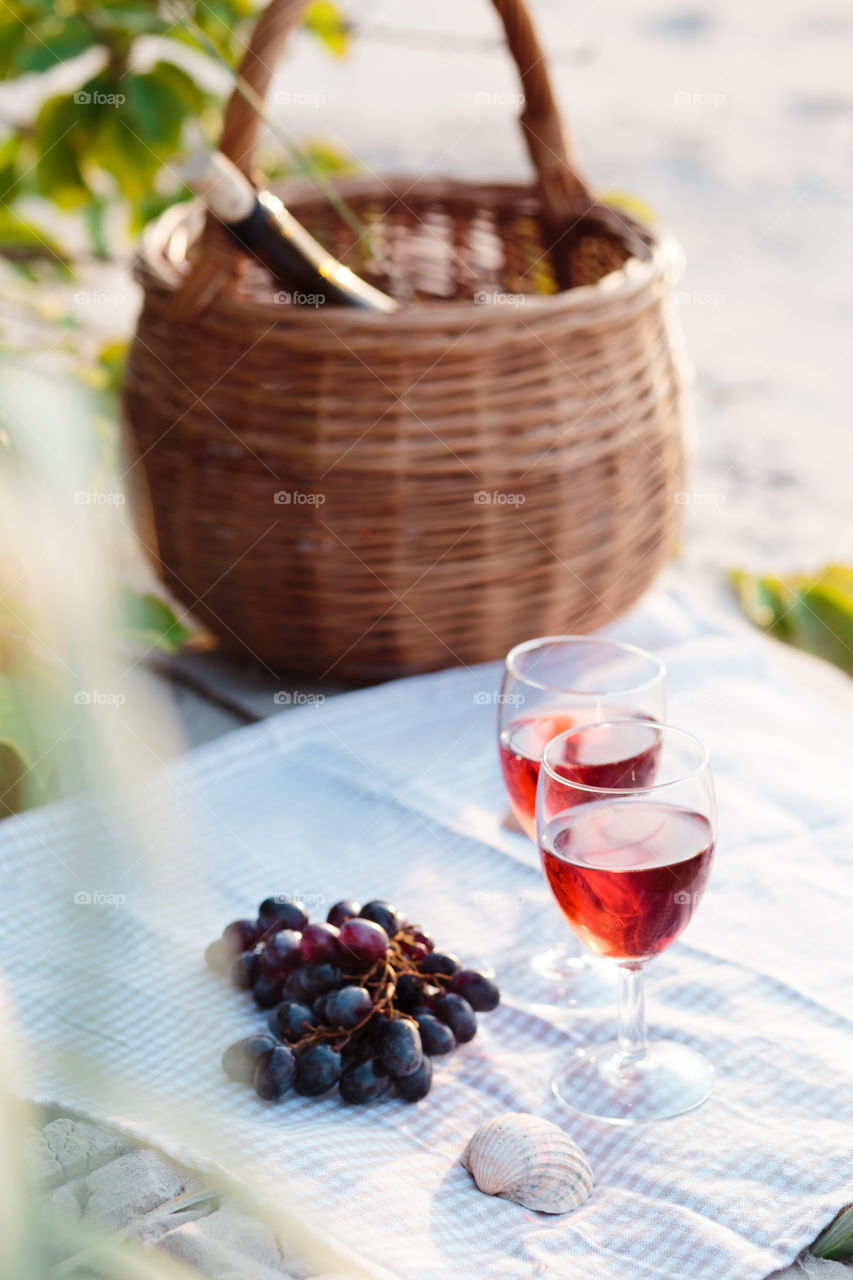 Two wine glasses with red wine standing on cloth, on beach, beside grapes and wicker basket with bottle of wine