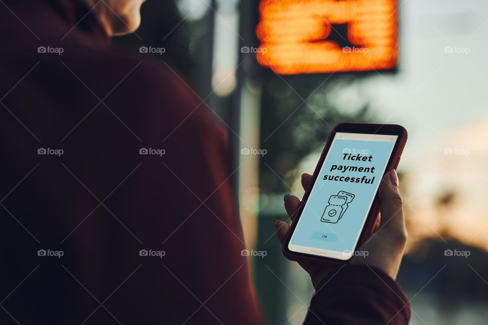Woman buying ticket online near bus stop before ride. Person paying for ticket using mobile payment app. Using technology on the go. Order confirmation on mobile screen. Top up account in app. Bus routes schedule in background