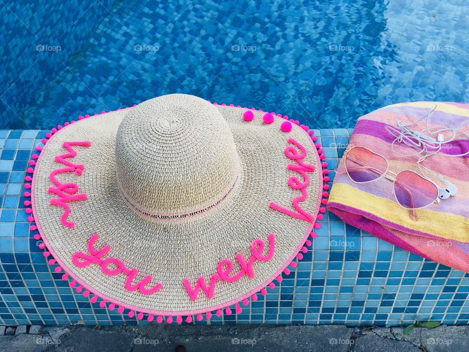 Summer hat with the words “wish you were here”,sunglasses, headphones and a pink towel on the edge of a pool with turquoise water