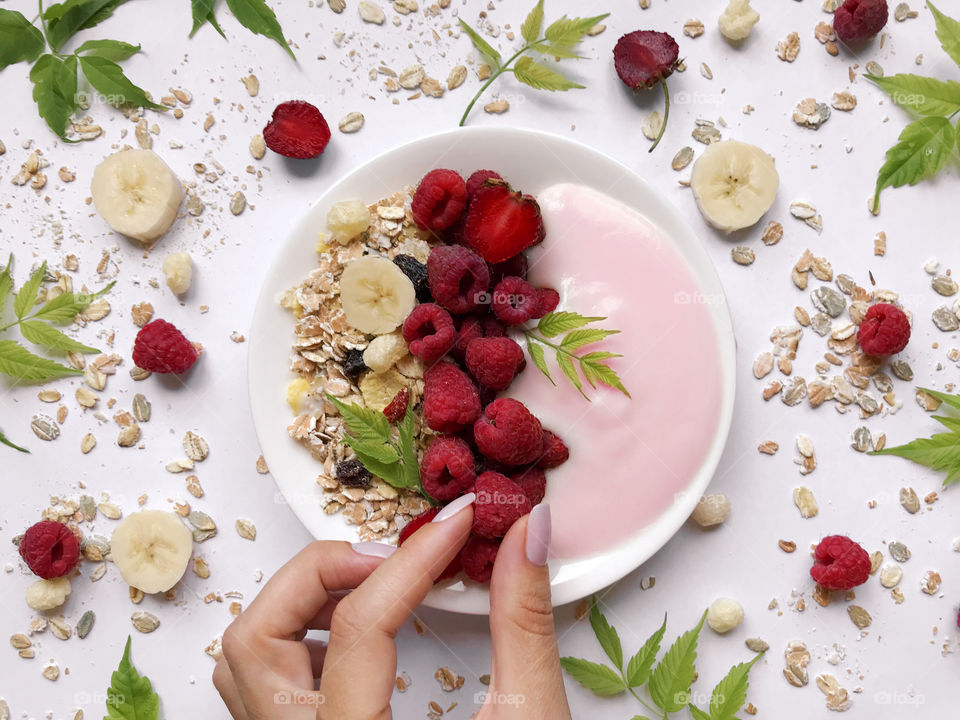 Female hand taking a red ripe raspberry from a breakfast bowl with yogurt, cereals and berries 