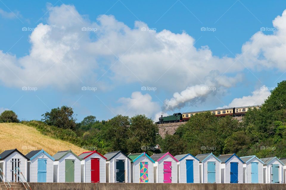 Colourful beach houses. Row of multi-colored beach huts with steam train puffing on stone arched viaduct against blue cloudy sky.