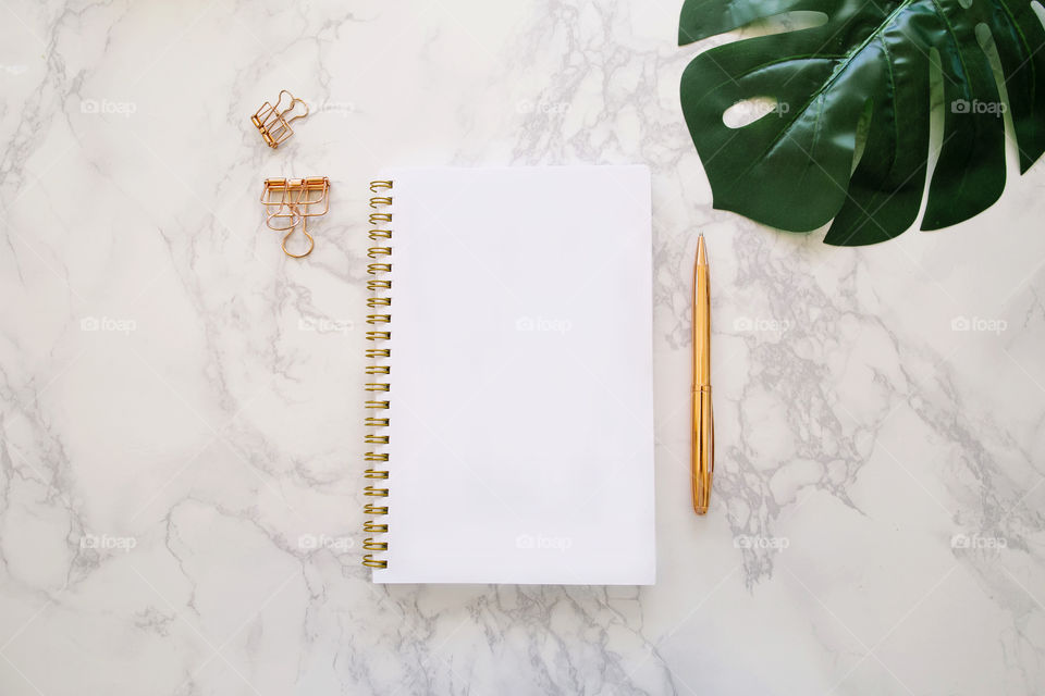 Modern marble workspace with rose gold pen and paper clips, notebook and monstera leaf; flat lay concept with copy space 