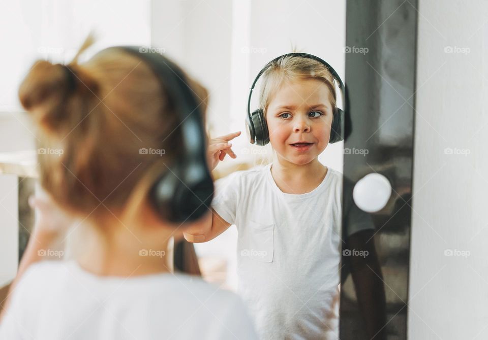 Cute toddler girl with fair hair in headphones looking in mirror