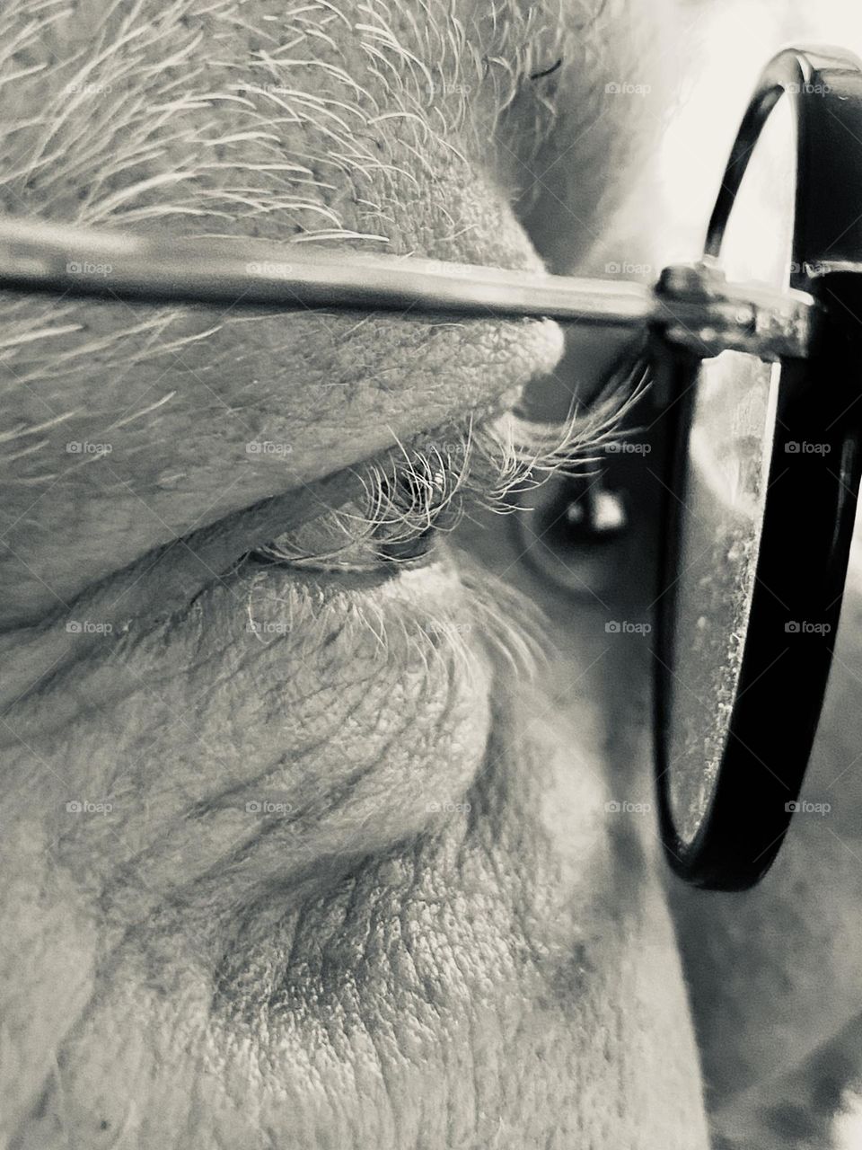 Close-up of an eye with black-framed, round glasses in front of it. You can see a small section of the face of a 55-year-old man, who is looking straight ahead. His wrinkles show his life experience. Photo in black / white.