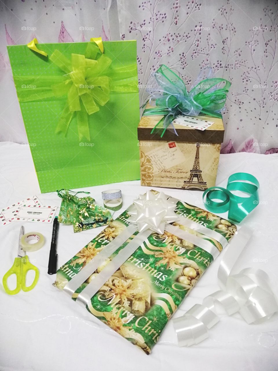 Packing Christmas presents showing some of the materials used. A green paper gift bag with green bow, a box with blue and green bow, and a present wrapped in a gift wrapper with Merry Christmas design and white ribbon.