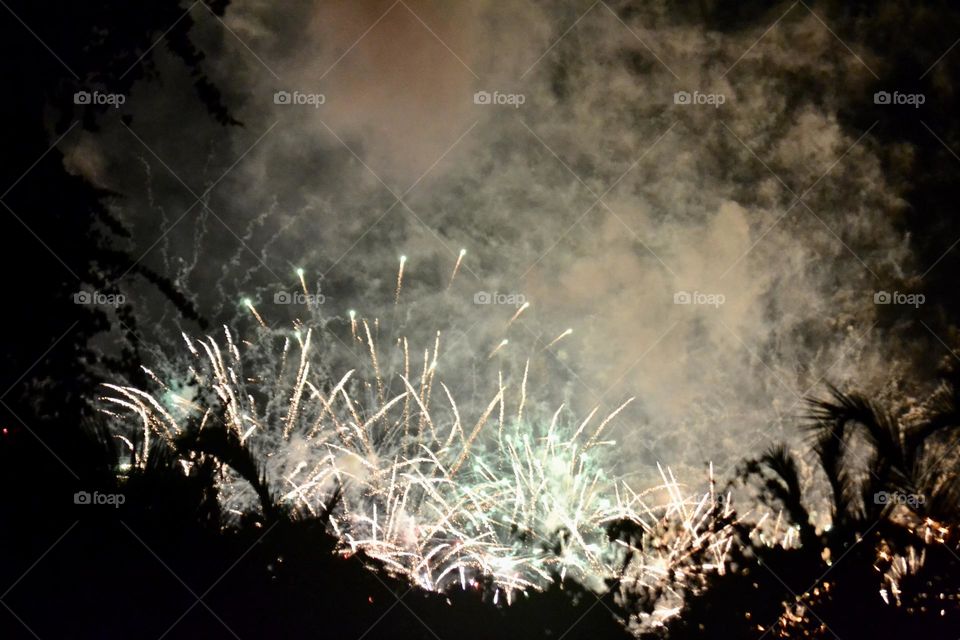 Bright white fireworks against a black smoke-filled sky in front of tropical palm trees