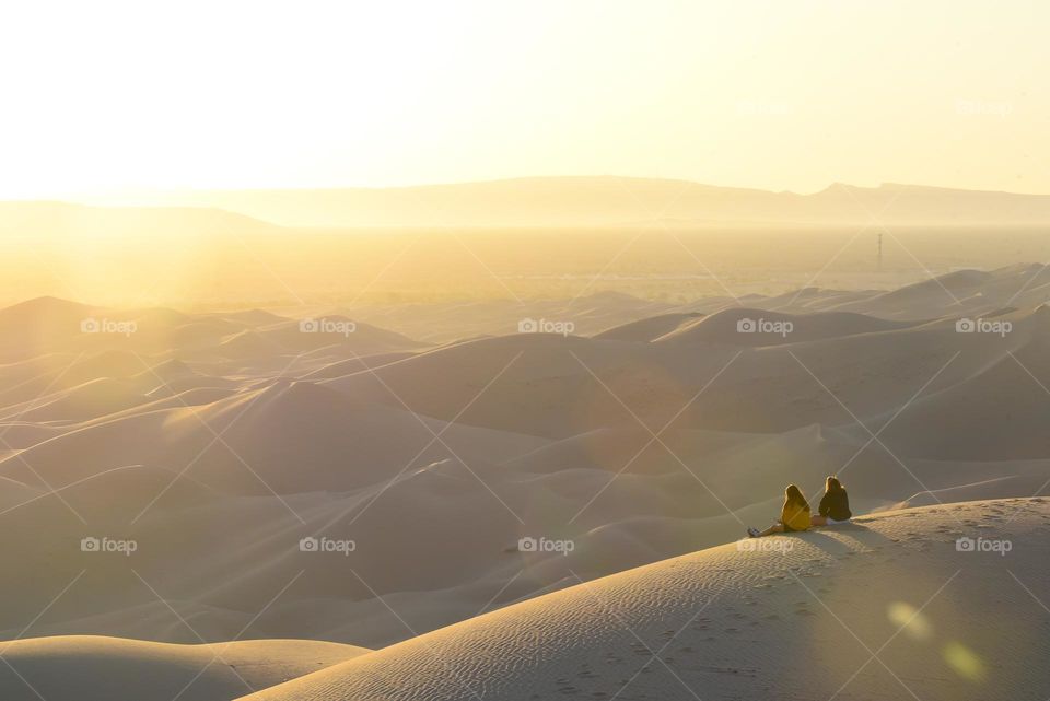 Two young people, friends, look in awe at the sunrise over the desert and admire the landscape and the color of the sun lighting up the sand dunes