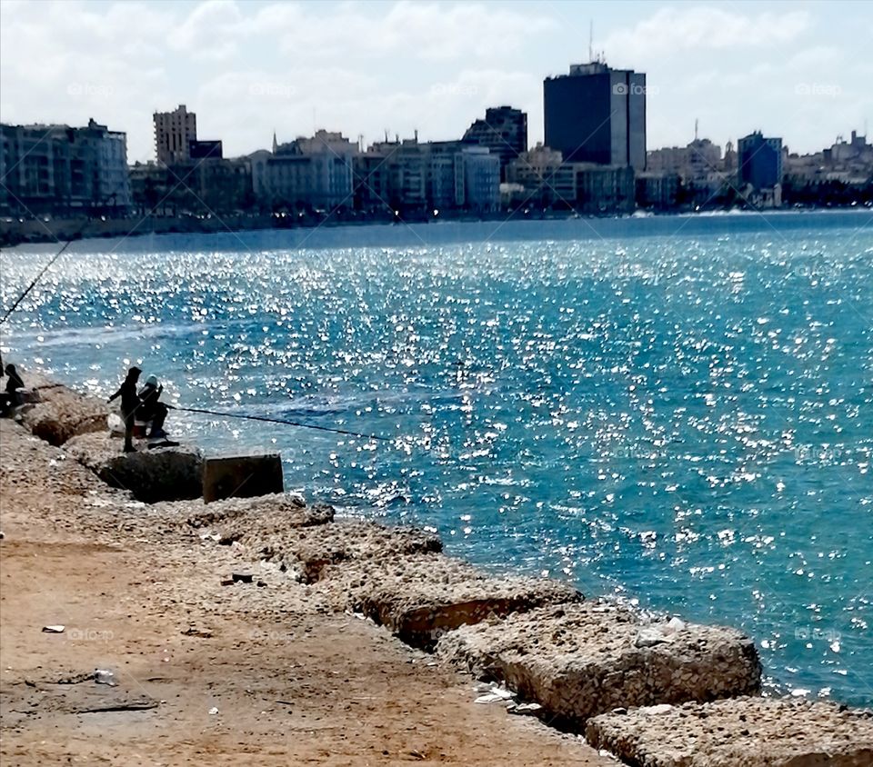 Some men practice a hobby fishing on the scenic Sea of Alexandria with apartment buildings, clear blue skies and beautiful clouds