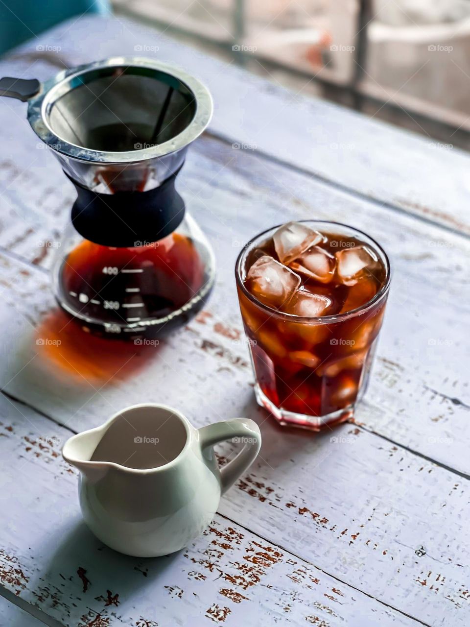 Iced black coffee on a white wooden table with small milk jug and coffee dripper