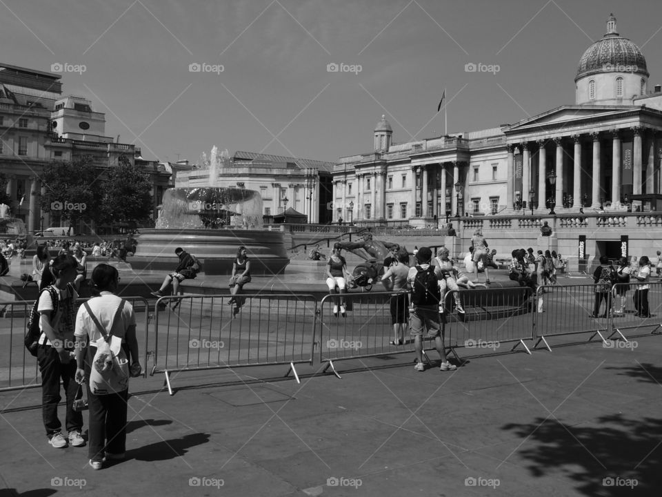 Crowds of people walk in an open square with a beautiful fountain in London on a sunny summer day. 