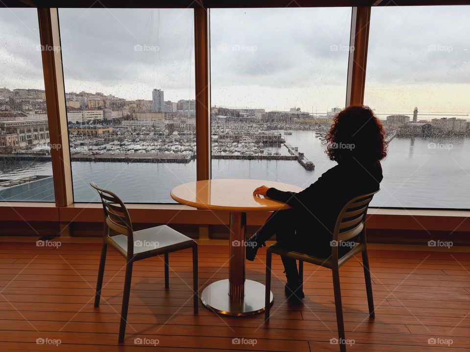 seen from behind, a woman sitting at a table looks out of a large window