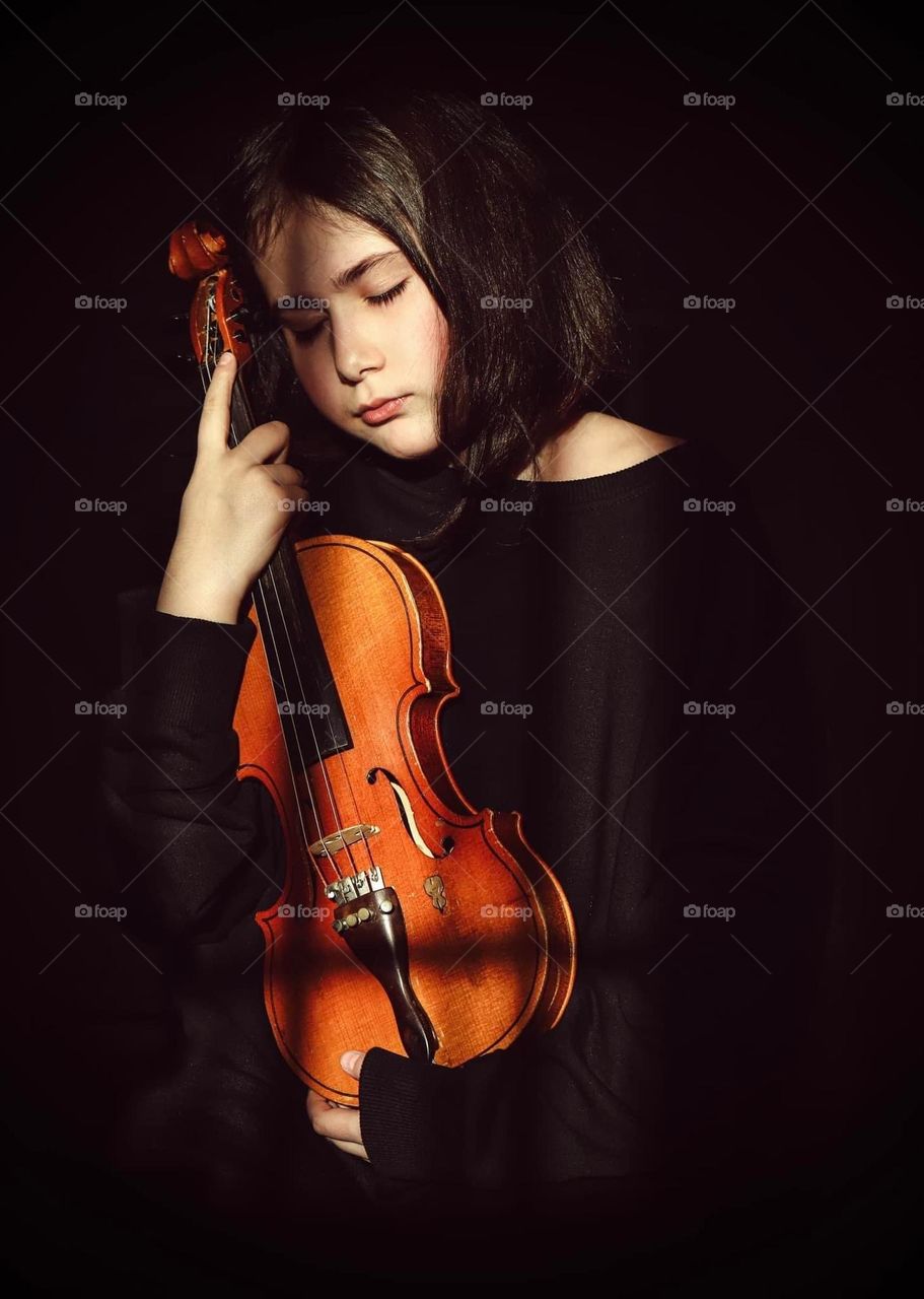 A little girl in a black dress holding a violin and a shadow from the windows falls on her