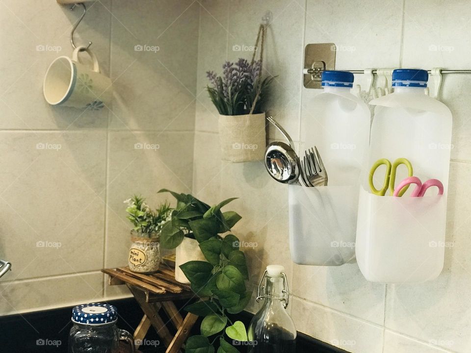 A plastic leftover milk bottles converted into a kitchen item’s holder like spoons , forks and scissors etc…and the surroundings covered with reusable coffee cup and glass bottles and containers and wooden stand eco friendly flower pots .