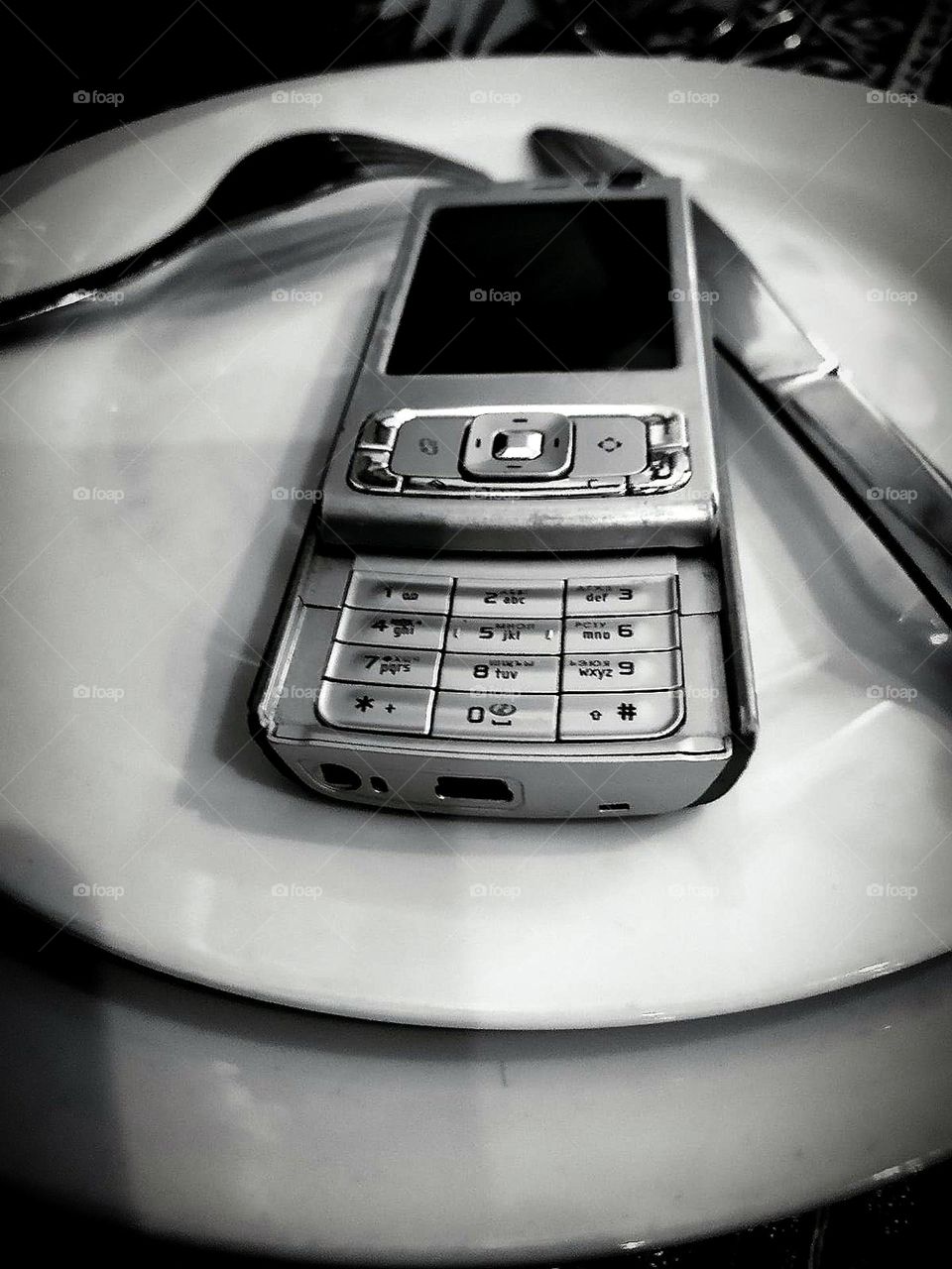 Black and white photo.  Contrast.  On the serving plates is an old push-button telephone: "Nokia 95".  Fork and knife are on the plates