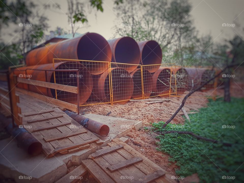 Giant rusty pipes are stacked on the ground behind the fence. Next to them is a wooden bridge.