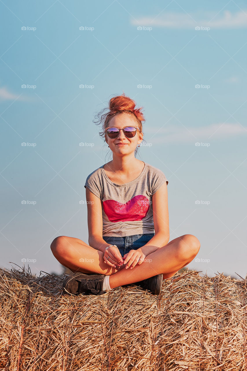 Happy smiling teenage girl sitting on a hay bale at sunset enjoying summer vacations in the countryside. Candid people, real moments, authentic situations