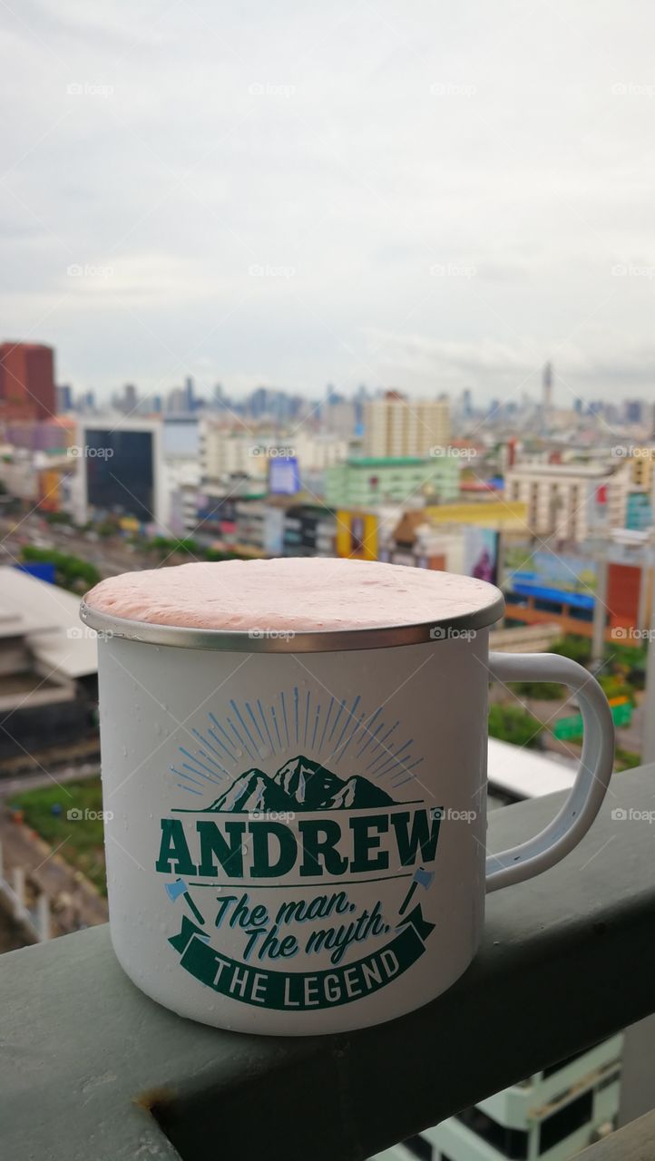 A strawberry smoothie on a balcony rail in Bangkok with a camping mug quoting Andrew is Legend.