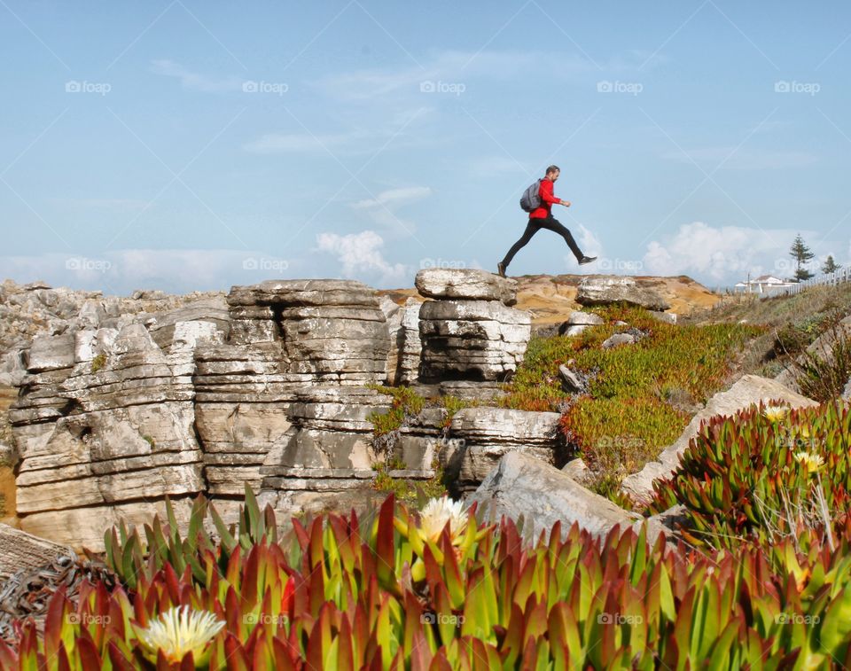 A man leaps over a gap between the rocks, there is a blue sky behind him and flowers in the foreground