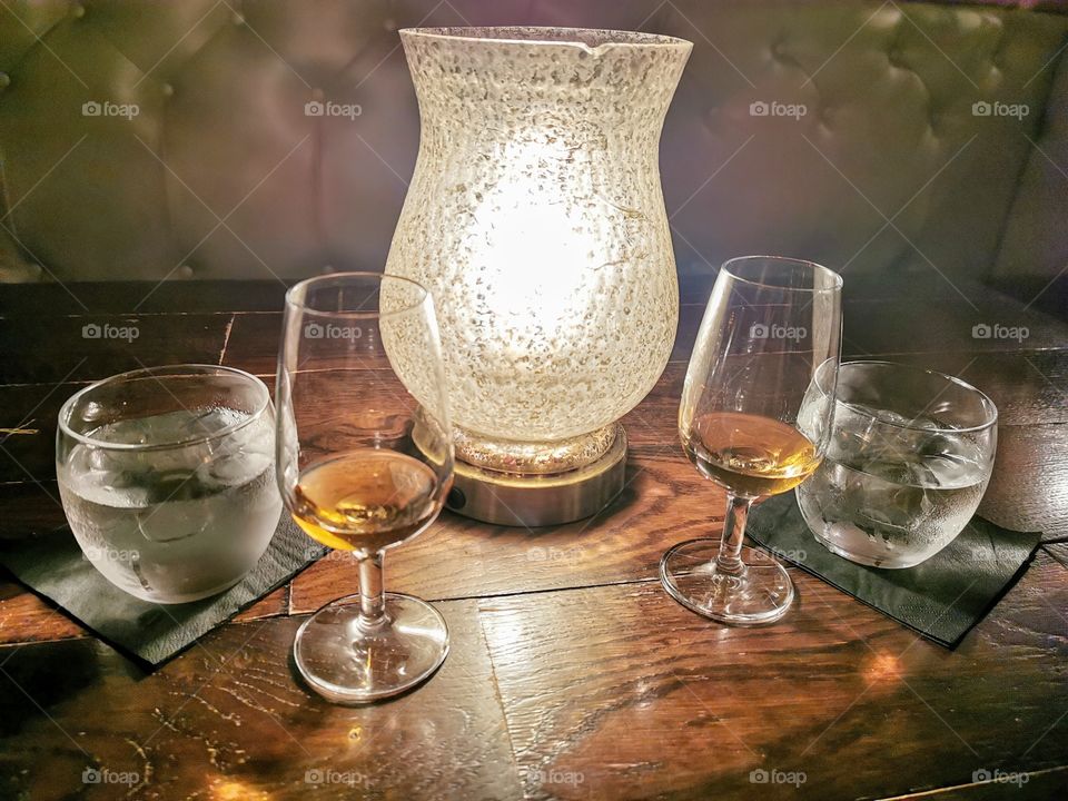 Two drams of whisky with two glasses of ice water either side of a table lamp on a wooden table with a leather bench in the background