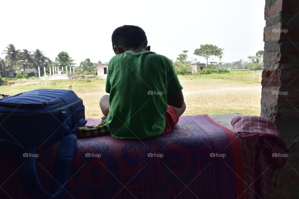 One little boy is sitting on a wall. The photo was taken from behind. The outside field is seen in the photo.