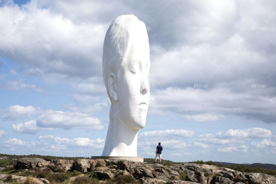 A man looking upon a giant woman’s head sculpture surrounded by clouds situated on the top of the rock 