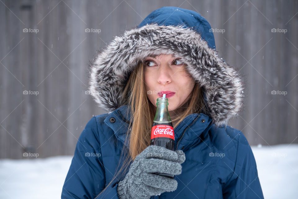 Millennial woman in a winter coat smiling and drinking a bottle of Coca-cola from a straw outdoors in the snow