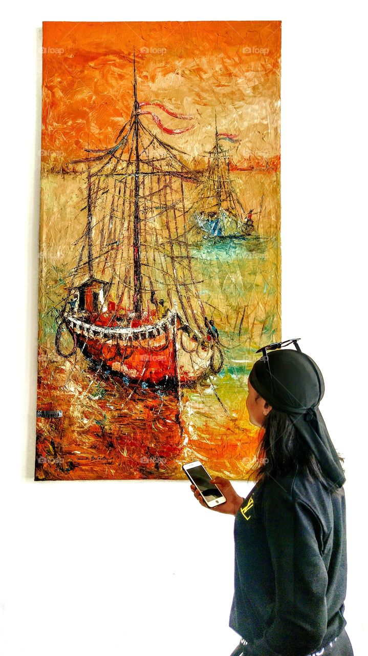 A lady holding an iphone and gazing at an artwork (painting) of a boat sailing in the sea