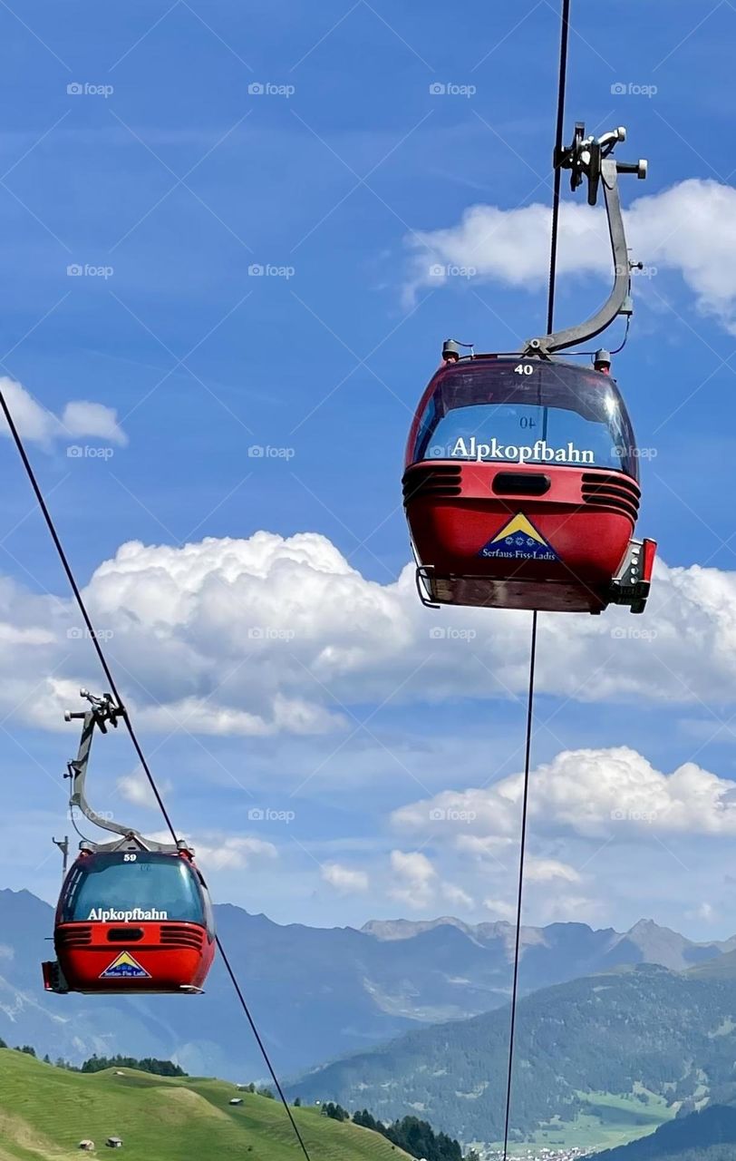Bright red gondolas pop against a vivid blue sky dotted with perfect white clouds, gliding above lush green ground and the majestic Austrian Alps in the distance.