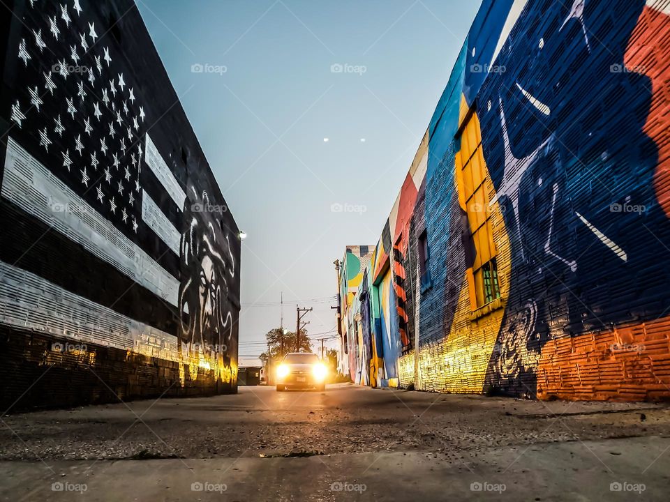 From the ground level,  a car driving through an alley at dusk and it's headlights are illuminating street art on both left and right exterior walls.