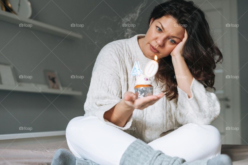 Young adult unhappy beautiful woman forty years plus size body positive in white cozy knitted sweater with festive cupcake with candle in hands on bed at home party, birthday girl