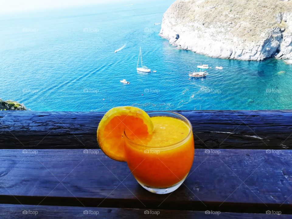the blue sea of ​​Ischia (Italy) is the background to a glass of freshly squeezed orange juice in the foreground