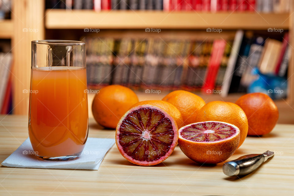 Glass of juice, knife and cut red oranges on a light wooden table. Blurred book shelves in the background.