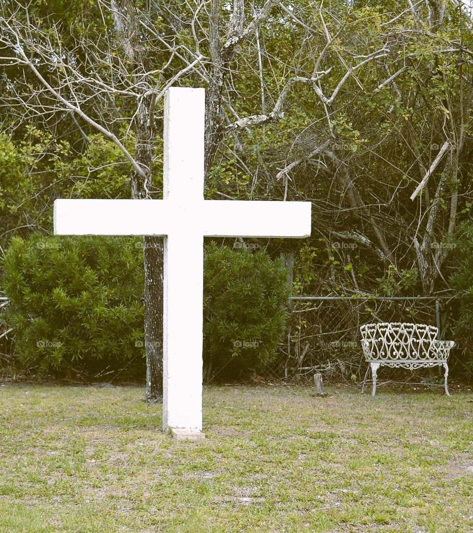 A large white cross above the grass and in front of trees and bushes next to an ornate iron bench