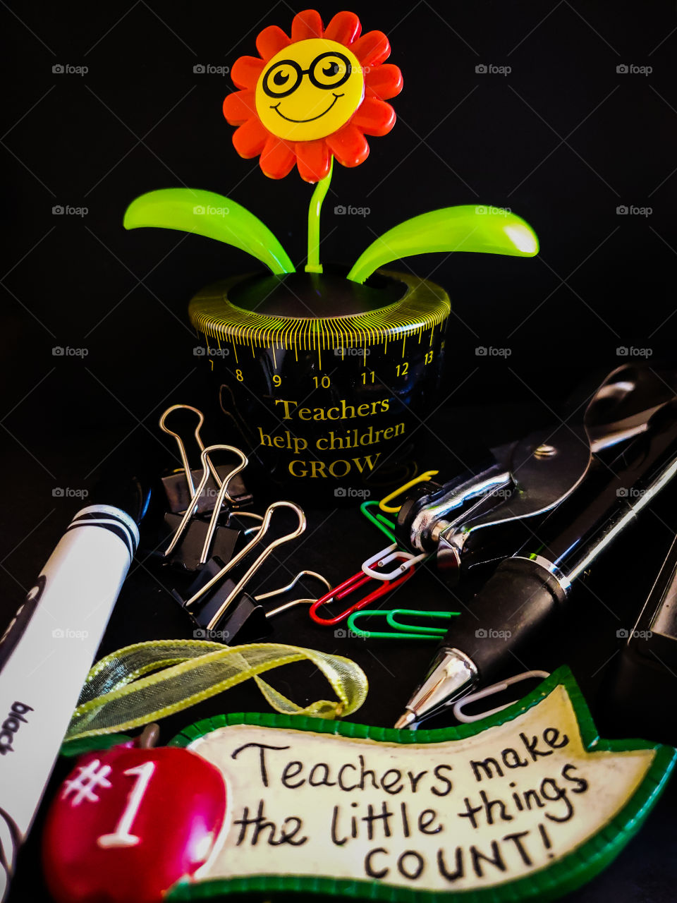 Still life of all things teachers/ educators including a solar plant that says teachers help children grow and an ornament that says teachers make the little things counts.  These are on a black background and surrounded my basic teacher supplies.