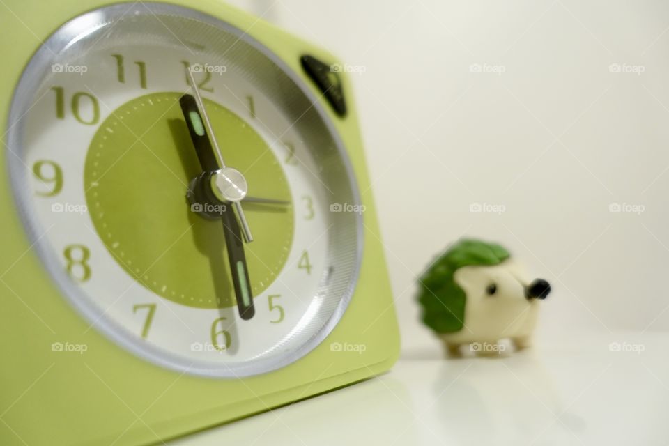 Green alarm clock with blurred hedgehog against white background.