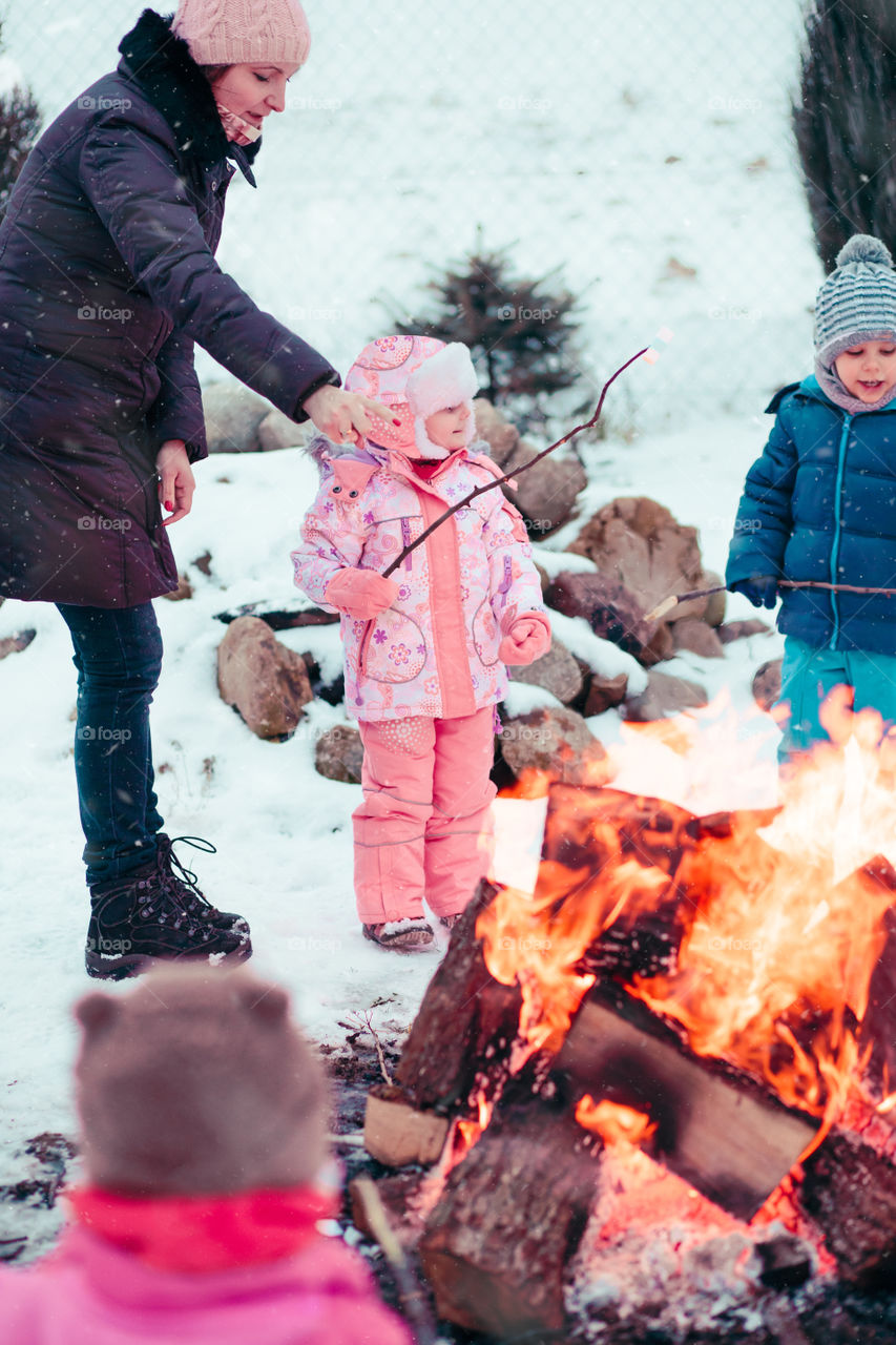 Family spending time together outdoors in the winter. Parents with children gathered around the campfire preparing marshmallows and snacks to toasting over the fire using wooden sticks